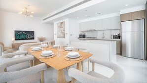 Ein moderner Essbereich mit einem Holztisch, gedeckt für sechs Personen, umgeben von grauen, gepolsterten Stühlen. Im Hintergrund ist eine Küche mit weißen Schränken und Edelstahlgeräten zu sehen.