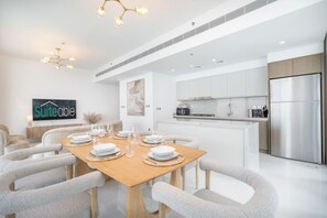 A modern dining area with a wooden table set for six, surrounded by grey upholstered chairs. A kitchen with white cabinets and stainless steel appliances is visible in the background.
