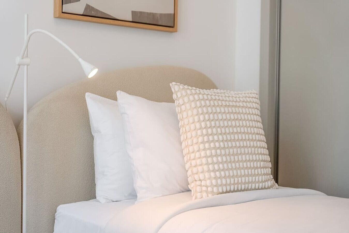 Una cama con ropa de cama blanca y dos almohadas, una lisa y otra con textura, en una habitación con una lámpara blanca y un cuadro en la pared.