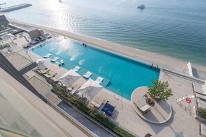 An aerial view of a hotel with a pool adjacent to a beach, a boat in the water, and a clear sky.