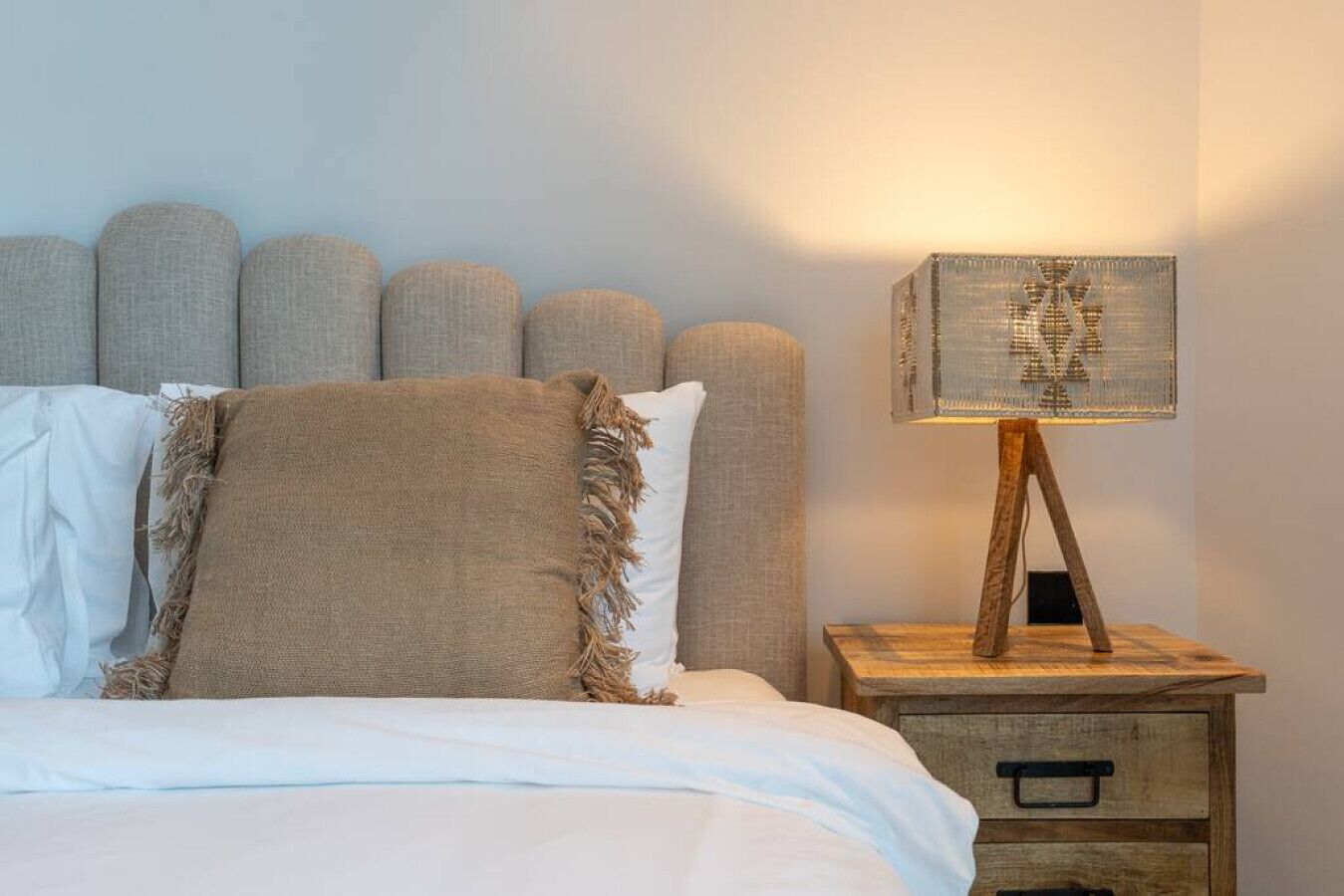 A bed with white bedding, a brown throw blanket, and a headboard with multiple cushions. There is a wooden nightstand with a lamp and a drawer.