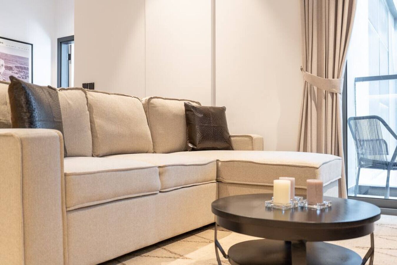 Un divano color beige con numerosi cuscini, un tavolino nero rotondo con candele e una finestra con tende.