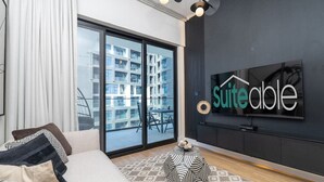 Une chambre d’hôtel moderne dotée d’une grande porte-fenêtre coulissante donnant sur un balcon, d’un canapé avec des coussins à motifs, d’un tapis rond et d’un téléviseur fixé au mur qui affiche le nom de l’hôtel.
