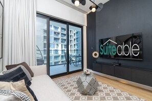 Une chambre d’hôtel moderne dotée d’une grande porte-fenêtre coulissante donnant sur un balcon, d’un canapé avec des coussins à motifs, d’un tapis rond et d’un téléviseur fixé au mur qui affiche le nom de l’hôtel.
