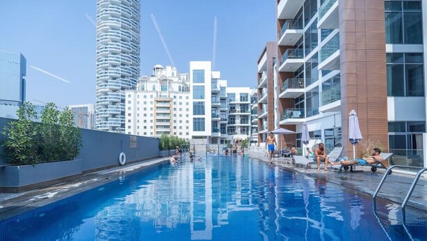 A swimming pool surrounded by modern buildings with balconies, a clear sky, and a few people relaxing by the pool. - Modern Comfort in a Spacious Apartment - Suiteable (دبي)