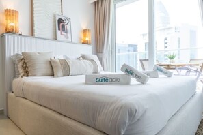 A neatly made bed with white bedding and pillows, a bedside table with a lamp, and a window with a view of buildings.