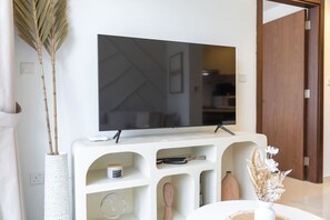 Ein modernes Wohnzimmer mit einem Flachbildfernseher an einer weißen Konsole, einer großen Vase mit Trockenpflanzen und einer Holztür im Hintergrund.