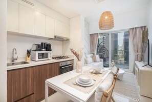 Eine moderne Küche mit weißen Schränken, einer Mikrowelle und einem Gasherd. Es gibt eine Essecke mit einem Tisch für zwei Personen, eine Couch und ein großes Fenster mit Blick auf die Stadt.