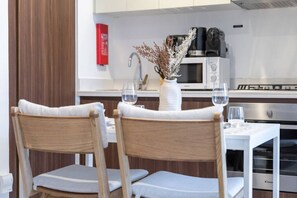 Ein Essbereich mit zwei Holzstühlen und einem weißen Tisch, eine Küche mit Mikrowelle und Kaffeemaschine sowie ein Feuerlöscher an der Wand.