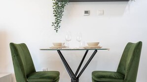 A dining area with a glass table, two green upholstered chairs, and a hanging plant. - Comfortable Broad Studio in Business Bay-Suiteable (Dubai)