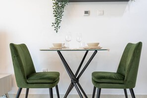 A dining area with a glass table, two green upholstered chairs, and a hanging plant.