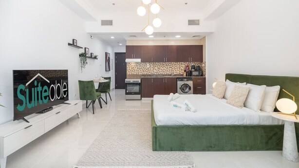 A modern hotel room with a green bed, a TV on a white stand, a kitchen area, and a dining space. - Comfortable Broad Studio in Business Bay-Suiteable (Dubai)