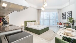 A modern hotel room with a kitchenette, a bed with green headboard, a dining area with a glass table, and a large window with curtains. - Comfortable Broad Studio in Business Bay-Suiteable (Dubai)