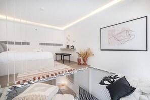 A modern bedroom with a bed, a desk, and a chair. There is a framed artwork on the wall and a small table with a vase of dried plants. - Fashionable loft with catamaran net in JVC (Dubai)