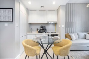 A modern kitchen with white cabinets, a gas stove, and a coffee maker. There is a glass dining table with two chairs in the foreground.