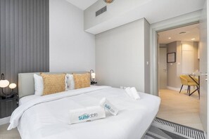 A modern hotel room with a neatly made bed, a bedside table with a lamp, and a small desk with a chair.