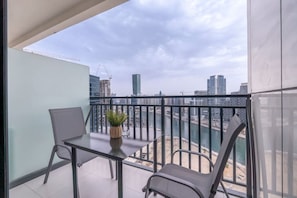 A balcony with a table and two chairs, a potted plant, and a view of a cityscape with buildings and a river.
