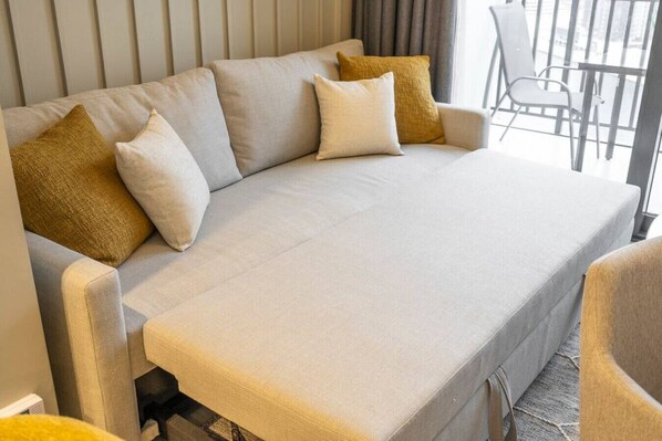 A sofa with a light-colored cover and two pillows, one beige and one mustard yellow, in a room with a window and a chair.