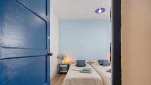 Una habitación con dos camas, una mesita de noche con lámpara y una puerta azul.