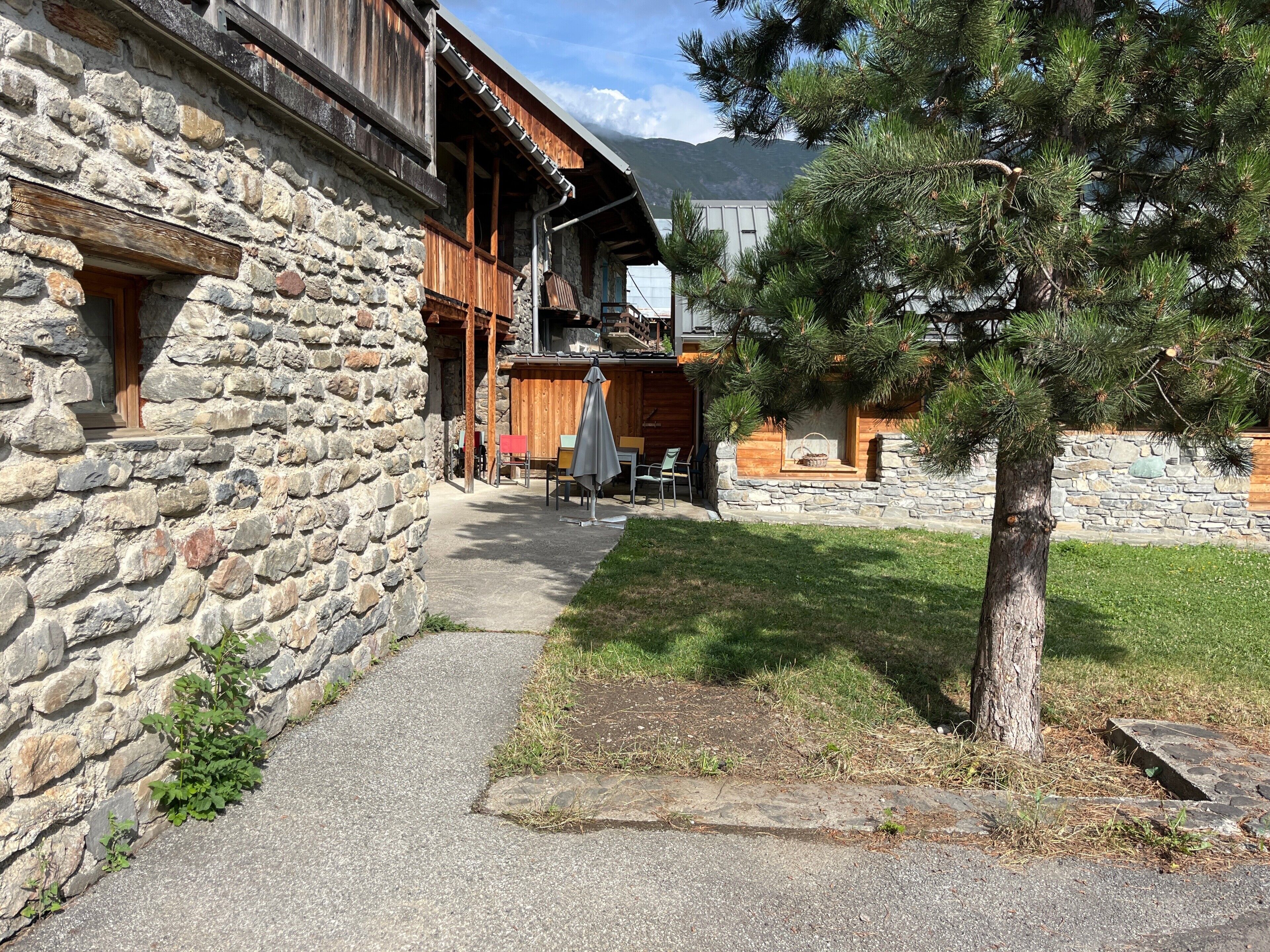 A stone building with a wooden balcony, a stone wall, and a grassy area with a tree.