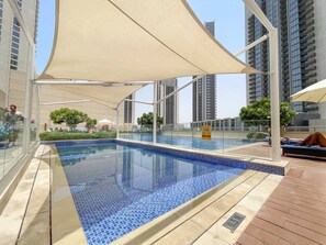 A rooftop swimming pool with a clear view of the city skyline, surrounded by modern buildings and a large canopy providing shade.