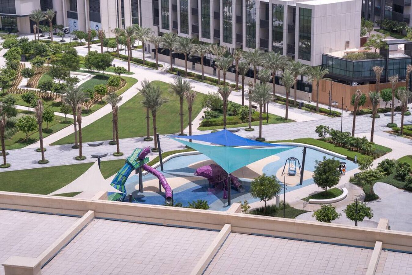 A modern outdoor area with a water play structure, palm trees, and a building with large windows.