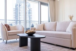 Ein modernes Wohnzimmer mit einer Couch, einem Sessel und einem runden Tisch mit einer Schale. Große Fenster bieten Blick auf hohe Gebäude.