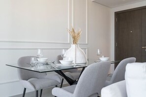 Et moderne spiseområde med et glasbord, grå polstrede stole, hvidt service og en vase med tørrede planter.