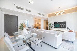 A modern living room with a glass dining table, white sofas, and a kitchen in the background.