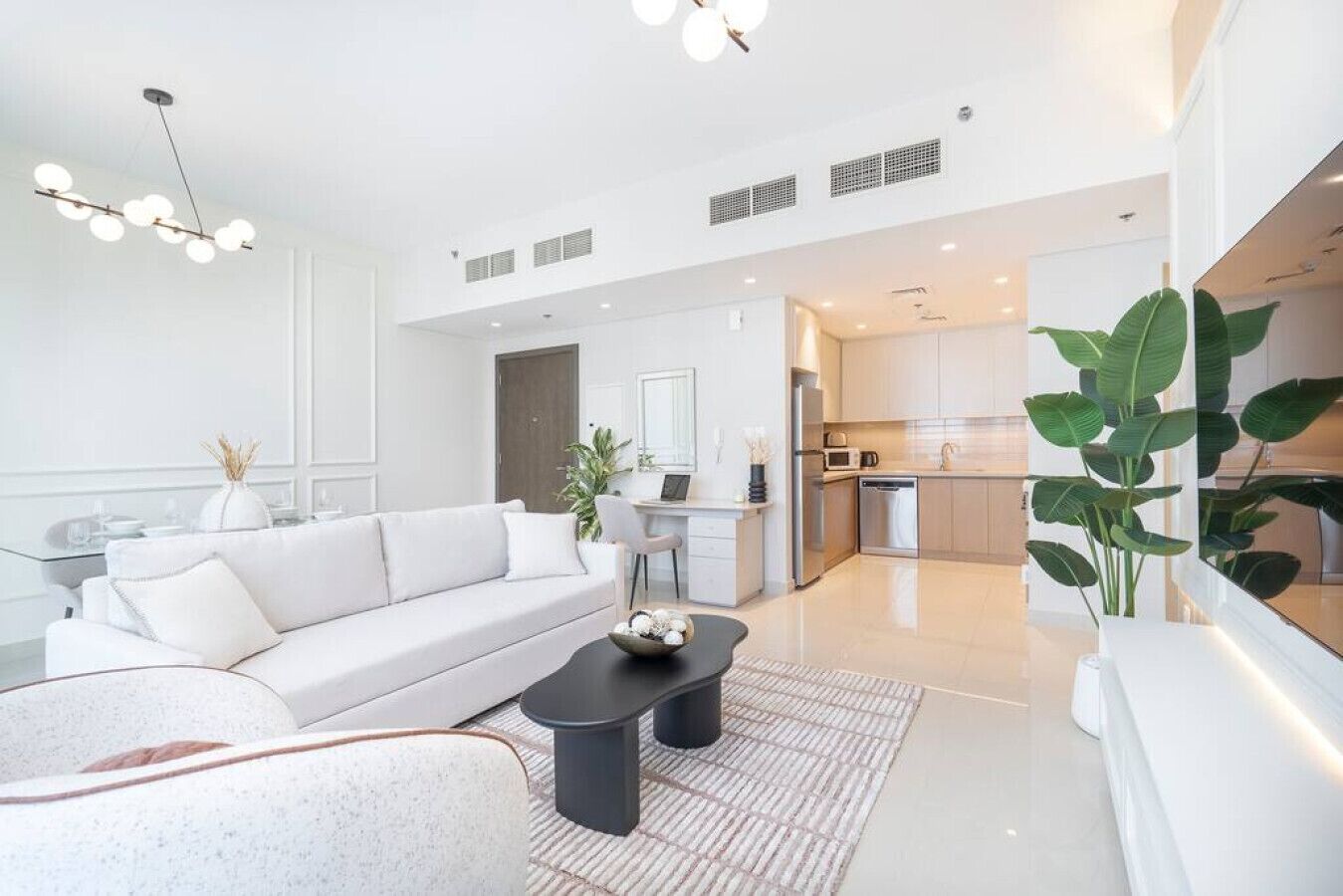 Ein modernes Wohnzimmer mit einem weißen Sofa, einem schwarzen Couchtisch und einem Essbereich mit einem weißen Tisch und Stühlen. Im Hintergrund ist eine Küche mit Edelstahlgeräten zu sehen.