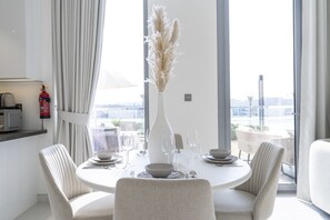 A dining area with a round table, white chairs, a vase with pampas grass, and a fire extinguisher on the wall.