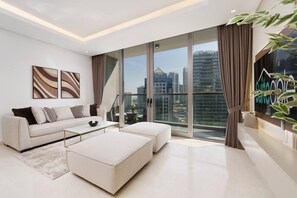 A modern living room with a sofa, ottoman, and coffee table, large windows with curtains, and artwork on the wall. - Business Bay Exquisite Living Experience-Suiteable (Dubai)