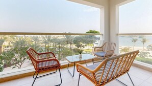 A balcony with wicker furniture, a glass table, and a view of a beach and palm trees.
