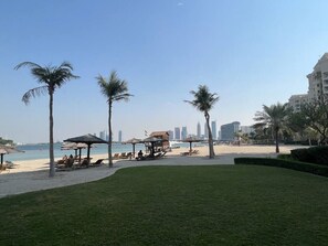 Une plage bordée de palmiers, avec des chaises longues et des parasols. On aperçoit des bâtiments au loin.