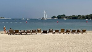 Ein Strand mit Liegestuhlreihen, die zum Meer ausgerichtet sind, ein Segelboot in der Ferne und eine kleine Insel mit Bäumen.
