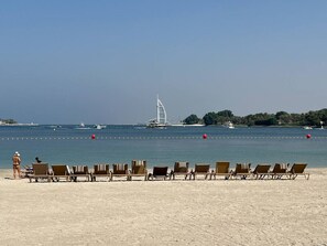 En strand med rækker af liggestole ud mod havet, en sejlbåd i det fjerne, og en lille ø med træer.