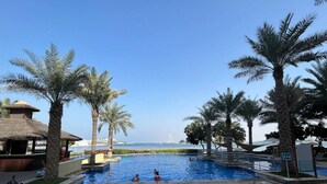 Ein Swimmingpool mit Palmen, ein Gebäude mit Reetdach und Blick aufs Meer.