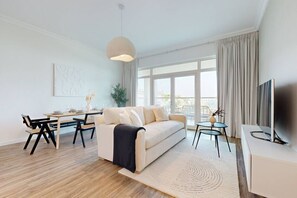 A modern living room with a sofa, dining table, and chairs, a TV, and large windows with curtains.