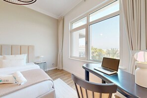 A hotel room with a bed, a desk with a laptop, a chair, a lamp, and a large window with curtains.