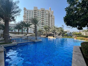 A large swimming pool with palm trees and a multi-story building in the background.