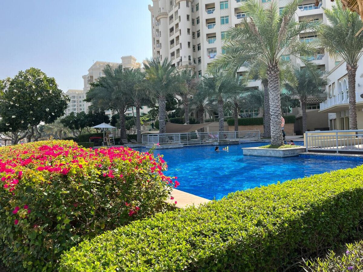 Una piscina rodeada de exuberante vegetación y palmeras, con un edificio de varios pisos al fondo.