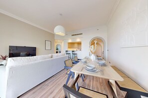 A modern living room with a sofa, dining table, and chairs. There is a TV and a kitchen area in the background.