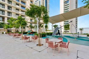 An outdoor patio area with tables and chairs, surrounded by greenery and a swimming pool.