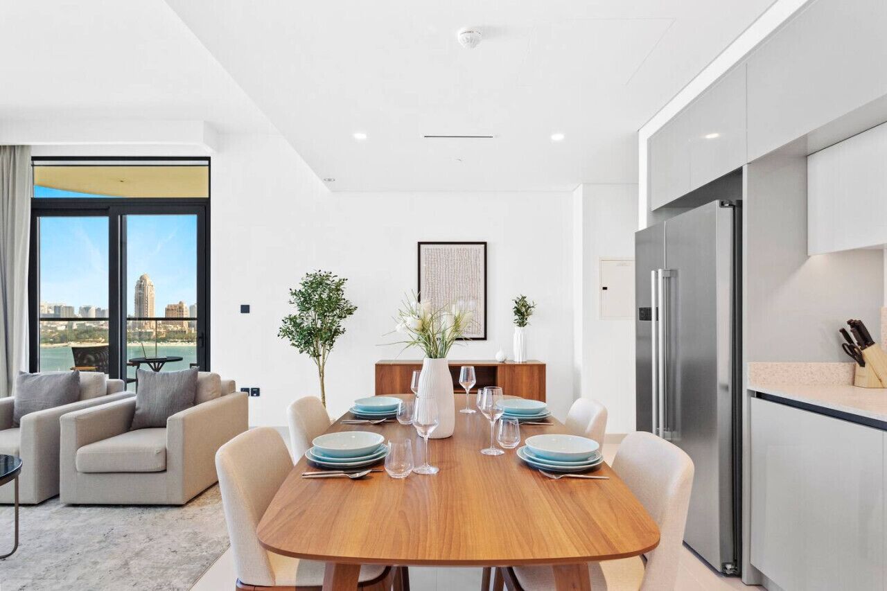 A modern dining area with a wooden table set for six, surrounded by chairs. A large window offers a view of a cityscape.