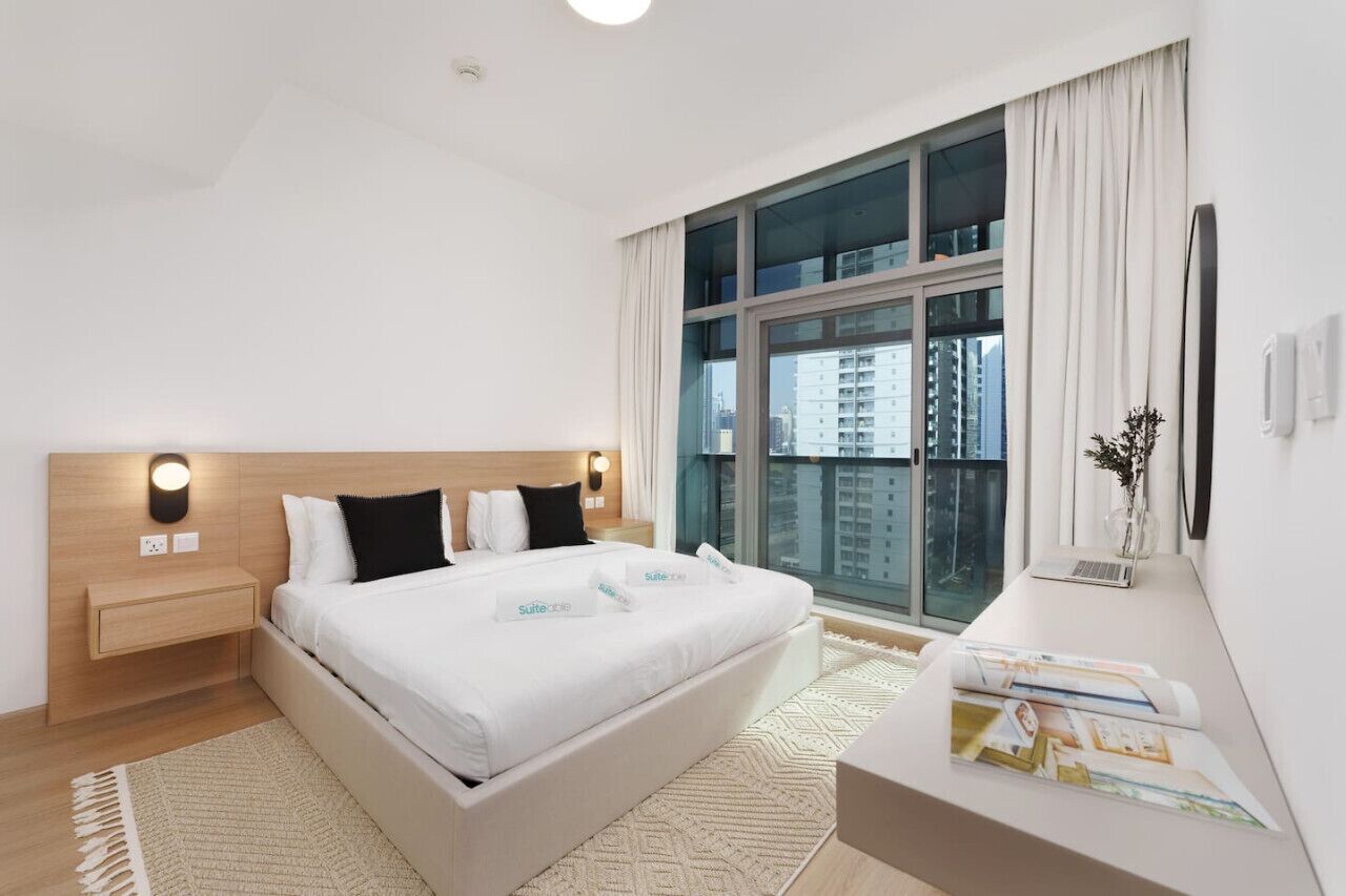 A modern hotel room with a large bed, a bedside table, a desk with a laptop, and a view of a cityscape through a large window.