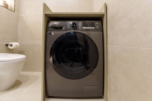 Un lave-linge compact et moderne, installé dans une salle de bain où se trouve également des toilettes, situées à sa gauche.