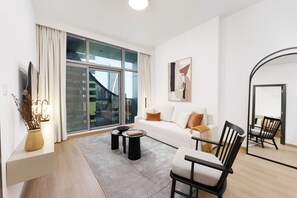 Un salon moderne avec un canapé, une table basse et des fauteuils, une grande fenêtre avec des rideaux et une œuvre d’art encadrée accrochée au mur.
