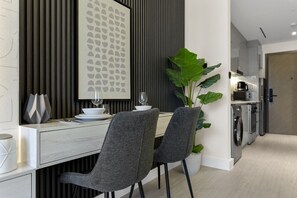 A modern kitchen with built-in appliances, a dining area with grey chairs and a white table, and a large green plant. - Studio Retreat with Canal and Burj Khalifa Views (Dubai)