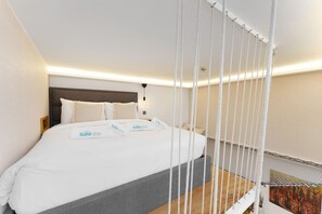 A hotel room with a bed, white bedding, a grey headboard, and a white curtain.