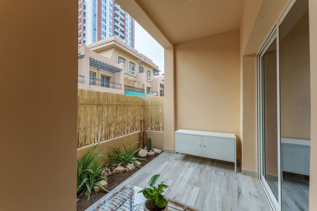 A balcony with a view of neighboring buildings, a small garden area with plants, and a white cabinet.